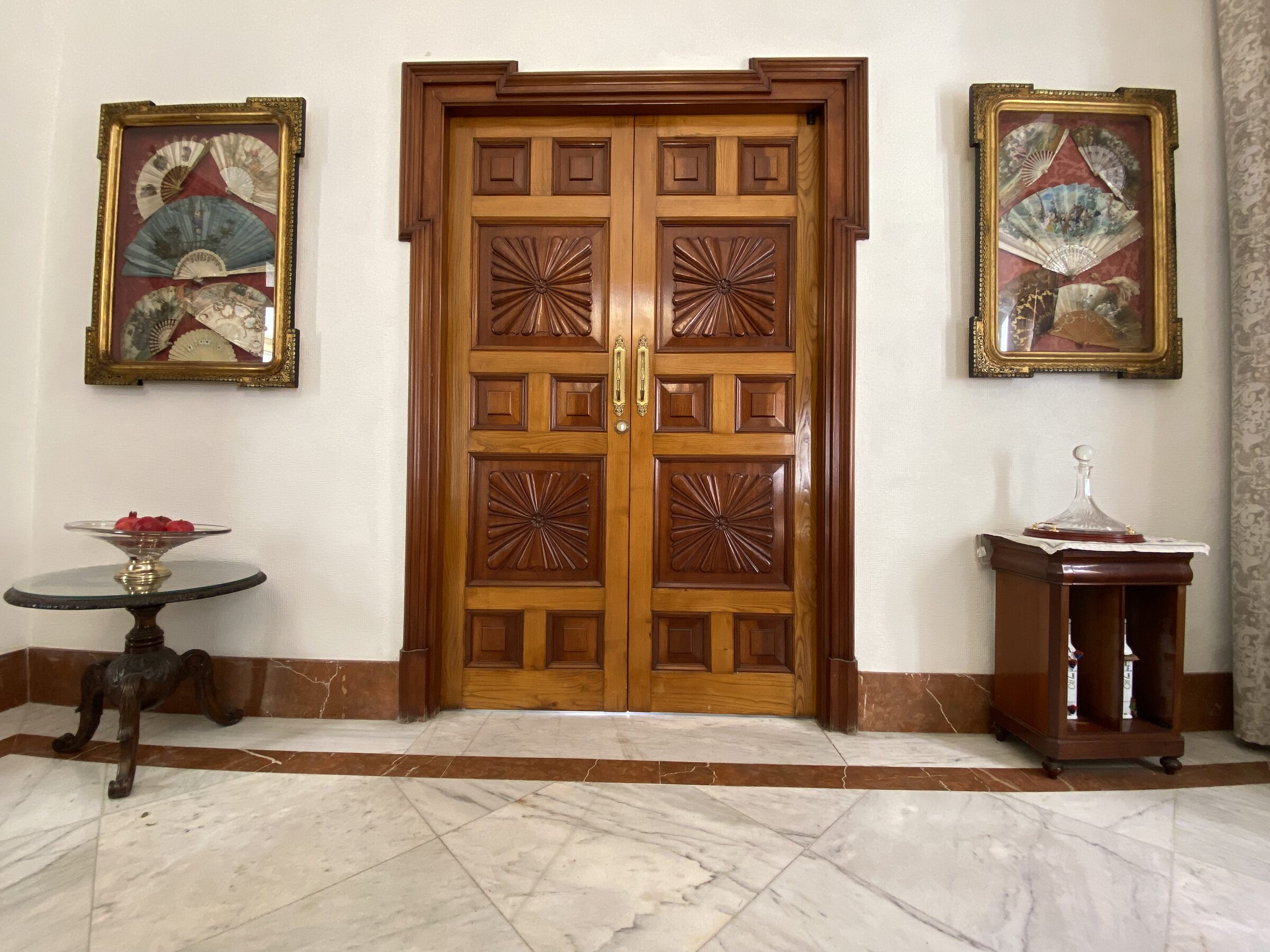 A deeply carved sun-burst-rosette door flanked by antique fans in gilt frames.