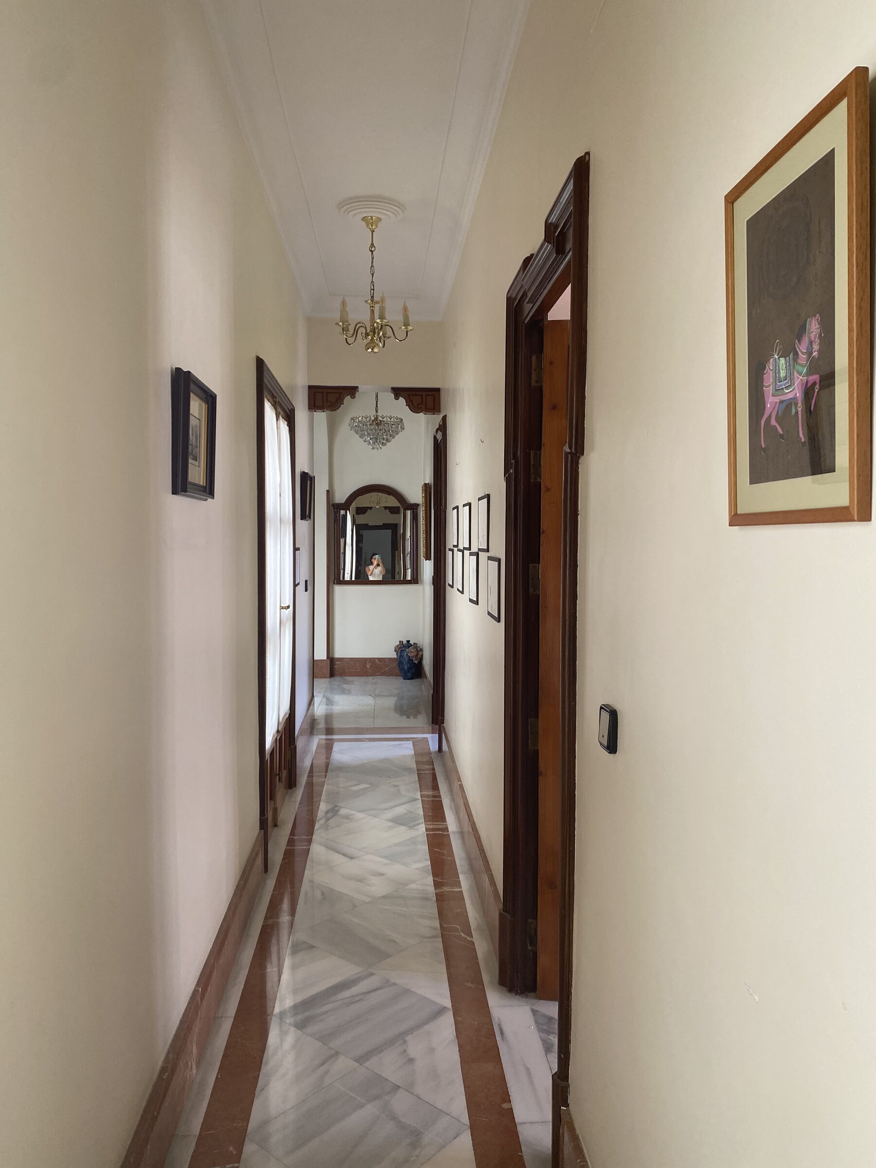 A corridor enfilade with a marble-strip floor, framed prints, a chandelier and a mirror at the far end.